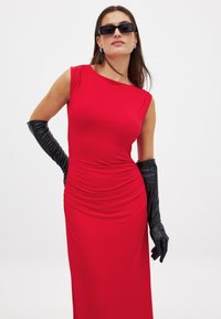 Czerwona sukienka bez rękawów o marszczonym kroju, wykonana z elastycznego materiału. Połączona z długimi czarnymi rękawiczkami i okularami przeciwsłonecznymi. Gładka tekstura.