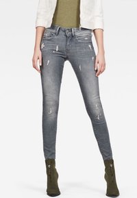 Jeans de mezclilla gris de corte ajustado, con ligero desgaste, detalles rasgados y un diseño de cinco bolsillos. Combinados con un suéter verde y botas de tobillo.