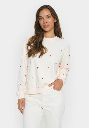 Cremfarbener Sweatshirt mit Rundhalsausschnitt, verziert mit kleinen roten Herzmustern und Rippbündchen. Kombiniert mit weißen Hosen.