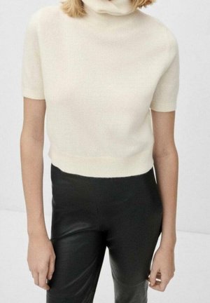 Femme portant un pull à col roulé crème à manches courtes associé à un pantalon en cuir noir taille haute, debout devant un fond uni.