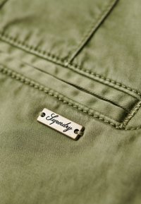 Tissu vert olive à texture lisse, avec une étiquette en métal gravée du mot 'Superdry', attachée près d'une couture de poche.