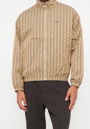 Beige jas met rits, geometrisch patroon en klein groen krokodillenlogo op de borst, gedragen met een donkere broek.