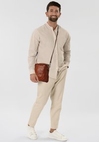 Borsa a tracolla in pelle marrone con texture liscia, forma rettangolare e tracolla regolabile. Indossata con una camicia beige chiaro con bottoni e pantaloni beige.