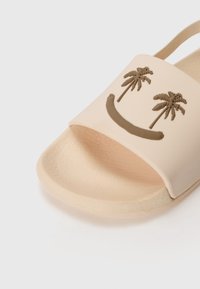 Infradito di colore beige chiaro con una soletta testurizzata. Presenta una fascia liscia con palme in rilievo e un sorriso. Materiale morbido e flessibile.