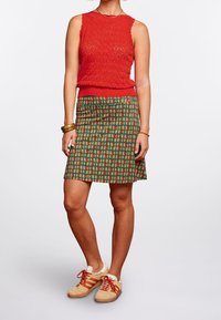 Röd stickad ärmlös tröja med ett texturerat mönster, tillsammans med en knä-lång grön kjol med ett färgglatt geometriskt tryck och beige sneakers.