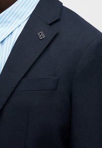 Blazer azul marino con un tejido texturizado, que presenta un bolsillo en el pecho y un pin de logo plateado en la solapa. Camisa de vestir a rayas azules debajo.