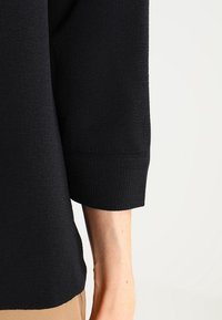 Schwarzes Langarmoberteil aus strukturiertem Stoff, mit rundem Halsausschnitt und entspannten Bündchen am Handgelenk, gezeigt an Schulter und Arm.