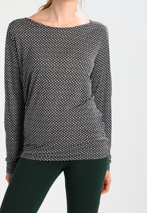 Femme portant un haut à manches longues noir et blanc à motifs avec un col bateau et un pantalon moulant vert foncé, debout devant un fond uni.