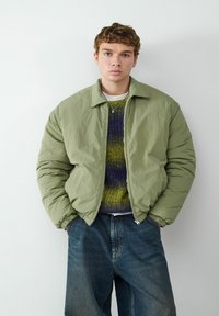 Jasnozielona bomberka z zamkiem błyskawicznym, ściągaczami na rękawach, nałożona na teksturowany sweter w odcieniach fioletu i zieleni, zestawiona z luźnymi niebieskimi dżinsami.