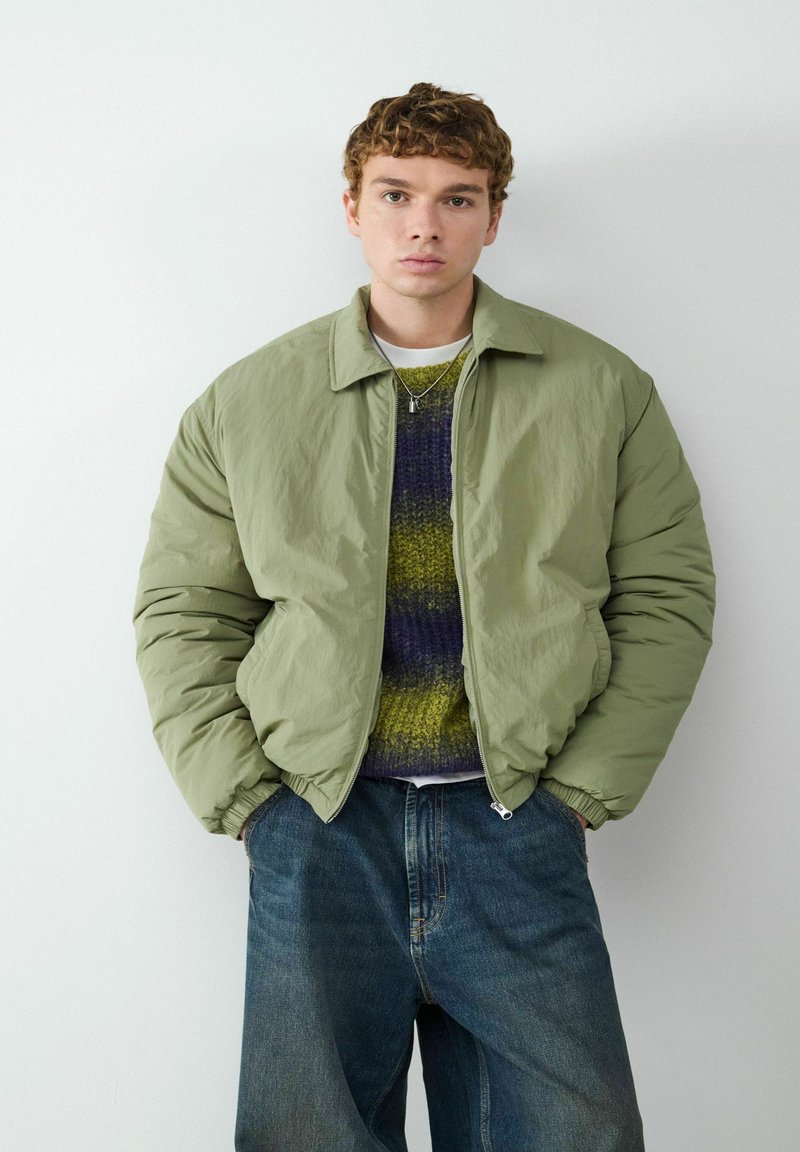 Jasnozielona bomberka z zamkiem błyskawicznym, ściągaczami na rękawach, nałożona na teksturowany sweter w odcieniach fioletu i zieleni, zestawiona z luźnymi niebieskimi dżinsami.