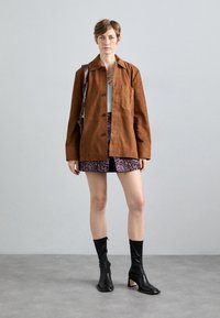 Γυναίκα που φοράει καφέ σακάκι με κουμπιά, φλοράλ φούστα, μαύρες μπότες μέσου ύψους και τσάντα ώμου, στέκεται σε γκρι δάπεδο μπροστά από ανοιχτό τοίχο.