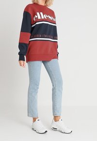 Czerwony i granatowy sweatshirt w stylu color-block z białym napisem, zestawiony z jasnoniebieskimi dżinsami i białymi trampkami. Gładki materiał, luźny krój.