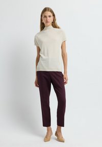 Lichtbeige top met korte mouwen en hoge halslijn, voorzien van textuuraccenten op de schouders, gecombineerd met dieppaarse op maat gemaakte pantalon en huidskleurige hakken.