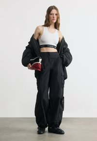 Γυναίκα με λευκό crop top, μαύρο φαρδύ σακάκι πέριξ των ώμων, μαύρο παντελόνι cargo, κρατώντας κόκκινη και λευκή τσάντα χειρός, στέκεται σε γκρι πάτωμα.