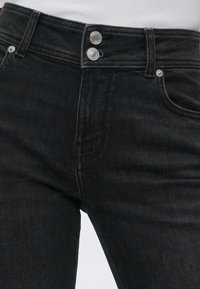 Κοντινή λήψη μέσης μαύρου τζιν jeans με διπλά ασημένια κουμπιά και θηλιές ζώνης, φορεμένα με άσπρο πουκάμισο μέσα στην μέση.