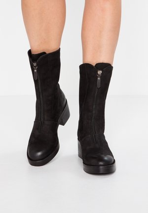 Czarne zamszowe botki do połowy łydki z przednimi zamkami, zaokrąglonymi czubkami i obcasami typu słupek, noszone przez osobę stojącą na białej podłodze.