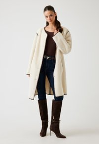 Cappotto di pile crema con collo a rever, rifinito con dettagli scuri. Abbinato a jeans scuri e stivaletti alti in suede marrone con tacco a spillo.