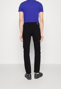 Osoba v modré košili s krátkým rukávem, černých džínách s viditelnou etiketou Calvin Klein a černých teniskách, stojící zády.