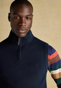 Mężczyzna w granatowym swetrze bez rękawów z zamkiem błyskawicznym, noszący kolorową, paskowaną koszulę z długim rękawem, na beżowym tle.