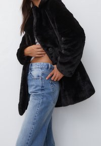 Zwarte faux bontjas met een zachte textuur, voorzien van een ronde kraag en twee knopen. Gecombineerd met high-waisted blauwe jeans.