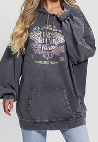 Femme portant un sweat à capuche noir délavé surdimensionné avec le texte « Los Angeles, Golden Road Tour 1987 », un graphisme ailé, une poche avant et un jean bleu clair.