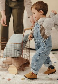 Bambino in salopette di jeans blu spinge un carrello di filo metallico con un cuscino blu chiaro e un oggetto a forma di stella color beige. Scarpe marroni con strap in velcro.