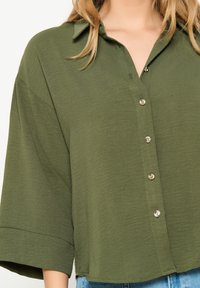 Olive green, short-sleeve button-up shirt with a collar, relaxed fit, and subtle texture. Features silver buttons along the front.