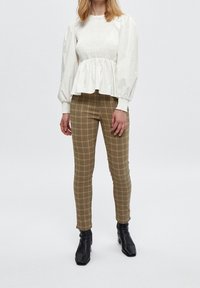 Bílý top s balónovými rukávy a nařaseným živůtkem, doladěný s obepnutými hnědými kostkovanými kalhotami. Celý outfit doplňují černé kotníkové boty.