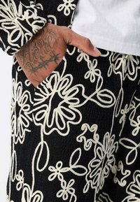 Main dans une poche de pantalon noir, ornée d'un tatouage de fil barbelé, avec des motifs floraux brodés en blanc et des motifs en boucle. L'ourlet de la chemise blanche est visible.