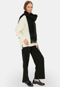Czarny szalik z frędzlami zarzucony na białym, oversize'owym swetrze, w zestawie z szerokimi czarnymi spodniami i błyszczącymi czarnymi butami.