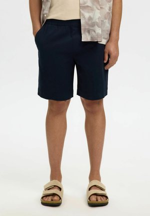 Mann trägt marineblaue knielange Shorts, beige offene Sandalen, beiges Hemd und ein gemustertes kurzärmeliges Überhemd, steht mit teilweise in die Taschen gesteckten Händen.