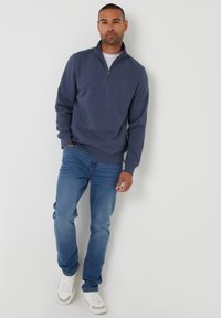 Sudadera azul marino con cremallera y puños acanalados, llevada sobre una camiseta blanca, combinada con jeans de color azul claro y zapatillas blancas; textura suave.