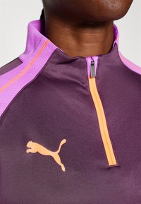Lilla atletisk kvart-zip topp med oransje glidelås, med lyse lilla detaljer og en fremtredende oransje Puma-logo foran.
