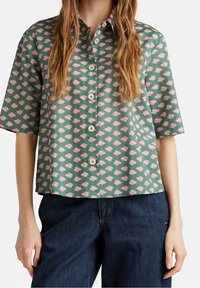 Donna che indossa una camicia verde a maniche corte con bottoni e motivo floreale rosa, abbinata a jeans blu scuro, in piedi contro uno sfondo bianco.