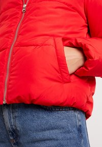 Red puffer jacket with a smooth finish, zip closure, and side pocket. Worn over blue denim, featuring a slightly cropped design.