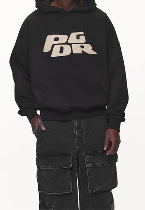 Mand iført sort hættetrøje med beige bogstaver "PGDR" og mørke cargobukser med store forlommer, stående mod en hvid baggrund.