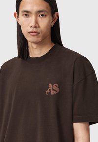 Brązowa bawełniana koszulka z zaokrąglonym dekoltem, krótkimi rękawami i subtelnym beżowym haftowanym logo "AS" na lewej piersi.