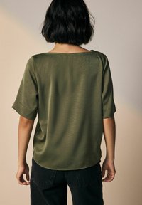 Top a maniche corte, verde oliva, realizzato in un tessuto leggero e texturizzato. Presenta un collo a barca e una vestibilità rilassata, con un orlo curvo.