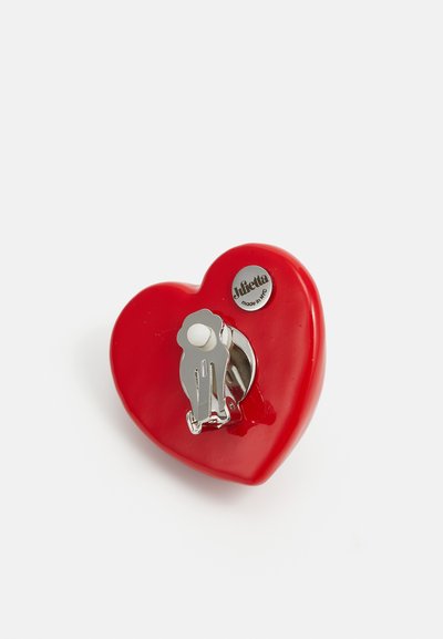Spilla a forma di cuore in plastica rossa lucida con una chiusura in metallo argentato. Presenta un piccolo design di scimmia argentata e un dettaglio di branding.