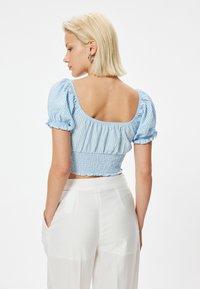Blau-weiße karierte, gekürzte Bluse mit Puffärmeln und Smock-Taille, kombiniert mit weißen, hochgeschnittenen Hosen. Glatte Stofftextur.