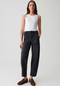 Pantaloni a vita alta in denim nero con una vestibilità rilassata, dotati di tasche frontali e cuciture a contrasto, abbinati a un top bianco senza maniche e scarpe marroni.