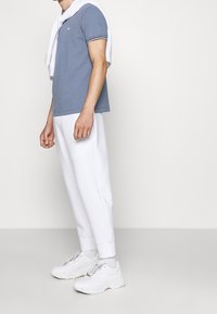 Polo de manga corta azul con detalles sutiles, sudadera blanca colocada sobre los hombros, combinada con pantalones de chándal blancos ajustados y zapatillas gruesas.