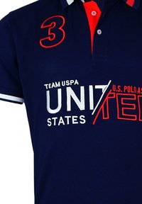 Cămașă polo bleumarin cu accente roșii. Conține logo-uri brodate "Team USA" și "U.S. Polo Assn." Include un guler roșu și un număr mare "3."