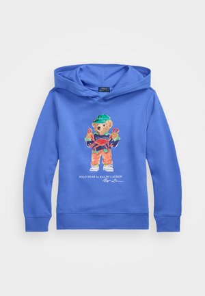 Plava dukserica s kapuljačom ima izvezeni grafički prikaz medvjeda u šarenoj odjeći, koji drži rakove, s tekstom "Polo Bear by Ralph Lauren" ispod.