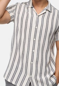 Mann trägt ein kurzärmliges, geknöpftes Hemd mit vertikalen schwarz-weißen Streifen, mit einer Hand erhoben in der Nähe des Kopfes.