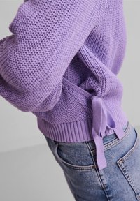 Fioletowy sweter z dzianiny o teksturowanym wzorze, z wiązanym detalem z tyłu oraz ściągaczami na mankietach i u dołu. Noszony z jasnoniebieskimi dżinsami.