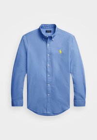 Světle modrá košile s dlouhým rukávem na knoflíky, vyrobená z bavlny. Má límec, zaoblený spodní okraj a malý žlutě vyšitý logo na hrudi.