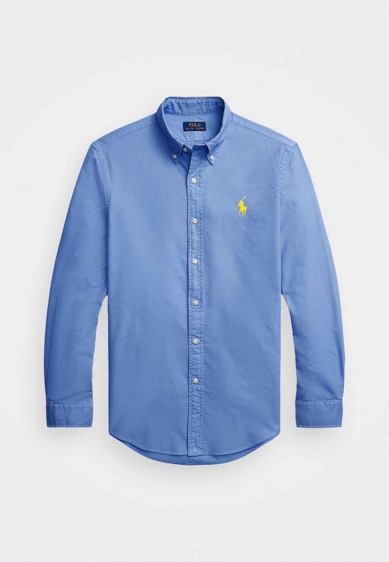 Světle modrá košile s dlouhým rukávem na knoflíky, vyrobená z bavlny. Má límec, zaoblený spodní okraj a malý žlutě vyšitý logo na hrudi.