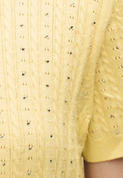 Κίτρινο πλεκτό ύφασμα με κάθετα σχέδια με καλώδιο και μικρές διασκορπισμένες ασημένιες στρας, που δείχνει μέρος ενός ενδύματος με κοντό μανίκι.