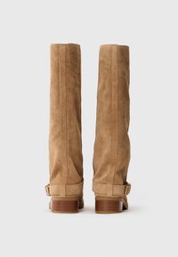 Un paio di stivali alti in suede beige con tacchi in legno impilati e cinturini con fibbie, mostrati da dietro su uno sfondo bianco.